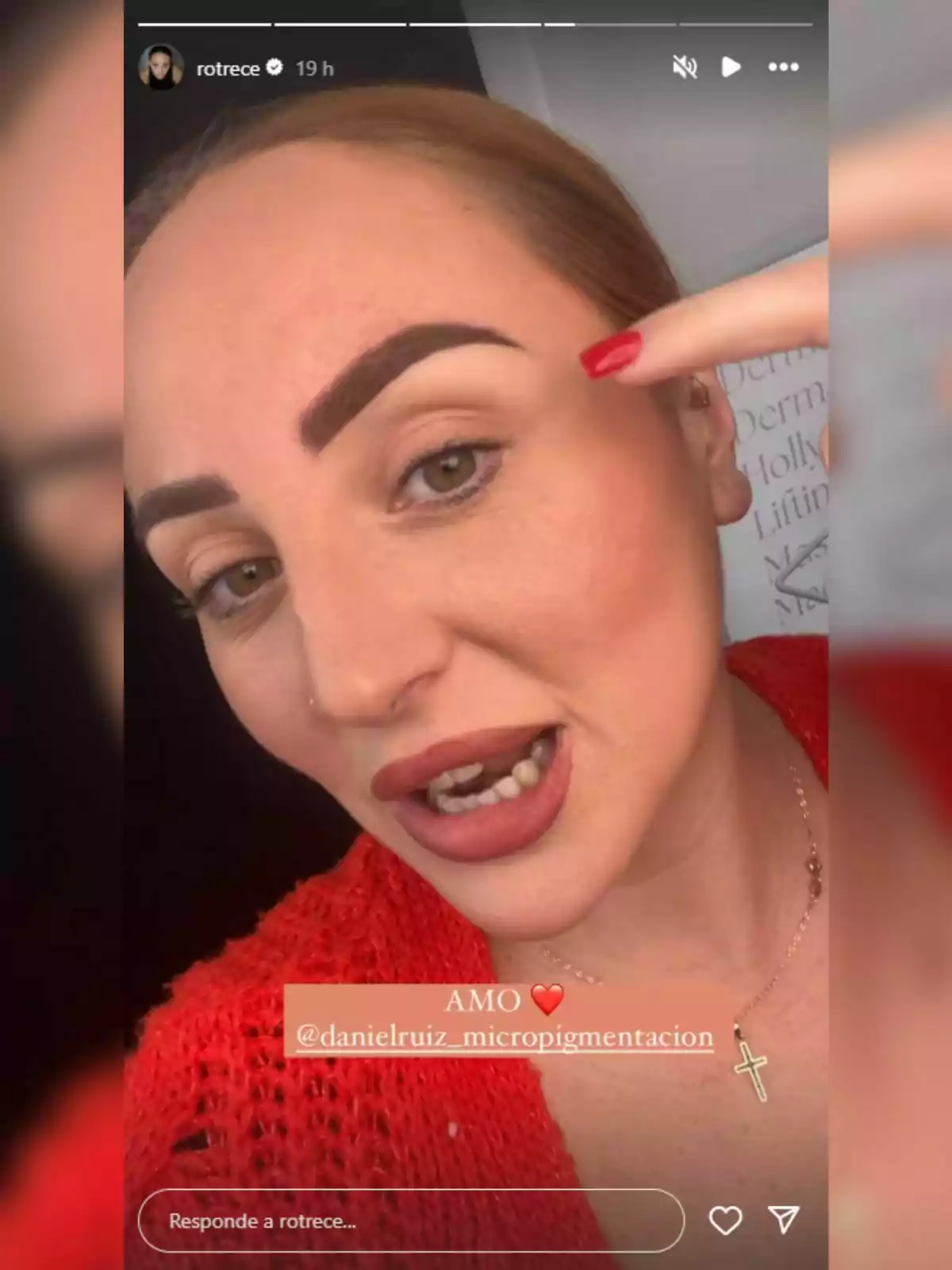 Una persona con maquillaje llamativo y cejas definidas señala su ceja mientras lleva un suéter rojo y un collar con un colgante de cruz. Una persona con maquillaje llamativo y cejas definidas señala su ceja mientras lleva un suéter rojo y un collar con un colgante de cruz.