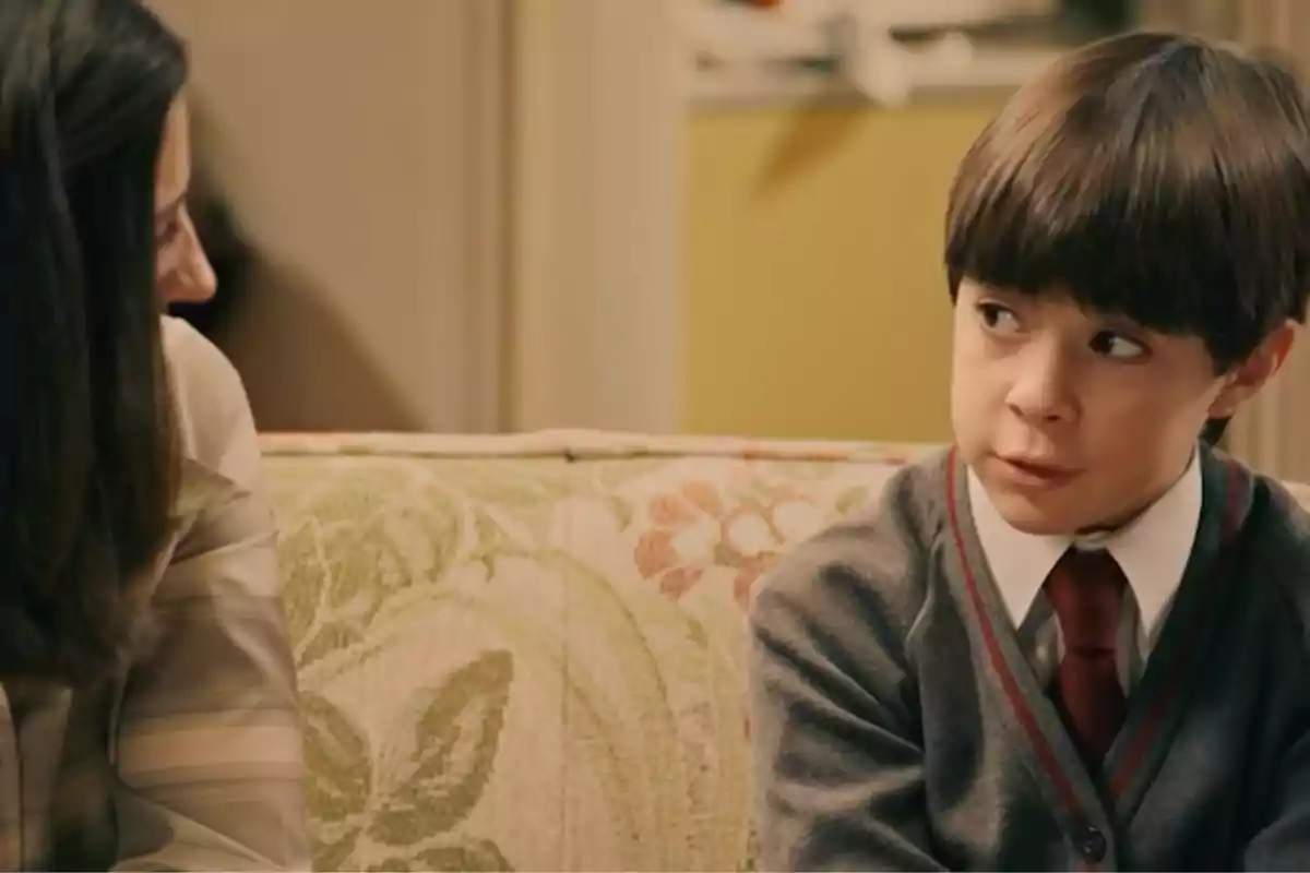 Teo con uniforme escolar y corbata roja está sentado en un sofá estampado junto a una mujer, ambos mirándose y conversando en una sala iluminada. Captura de 'Sueños de libertad' Teo con uniforme escolar y corbata roja está sentado en un sofá estampado junto a una mujer, ambos mirándose y conversando en una sala iluminada. Captura de 'Sueños de libertad'