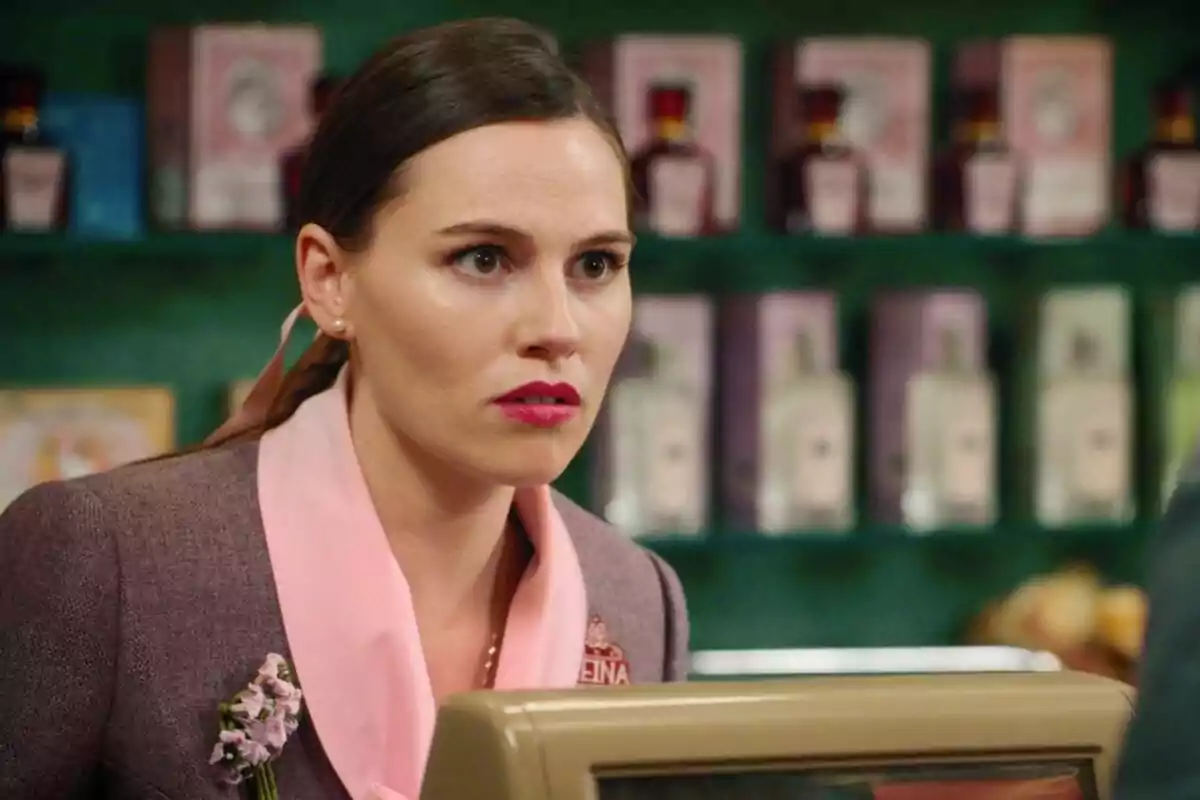 Claudia con expresión seria y labios rojos está detrás de un mostrador en una tienda, con estantes llenos de productos en el fondo. Captura de 'Sueños de libertad' el 21 de mayo de 2024