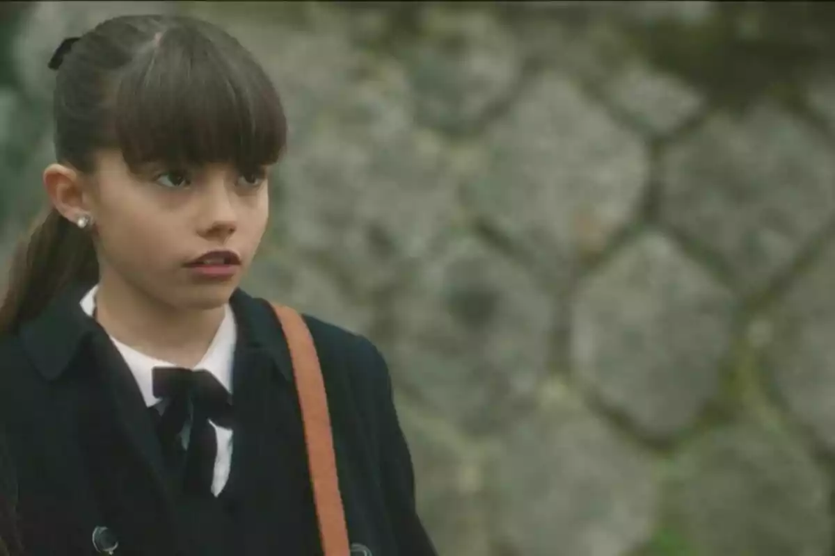 Julia con uniforme escolar y expresión seria, con un fondo de pared de piedra. Captura de 'Sueños de libertad'