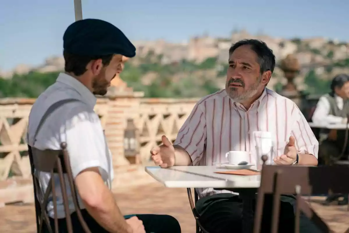 Luis y Alberto conversan en una terraza al aire libre con una ciudad desenfocada al fondo. Captura de 'Sueños de libertad'