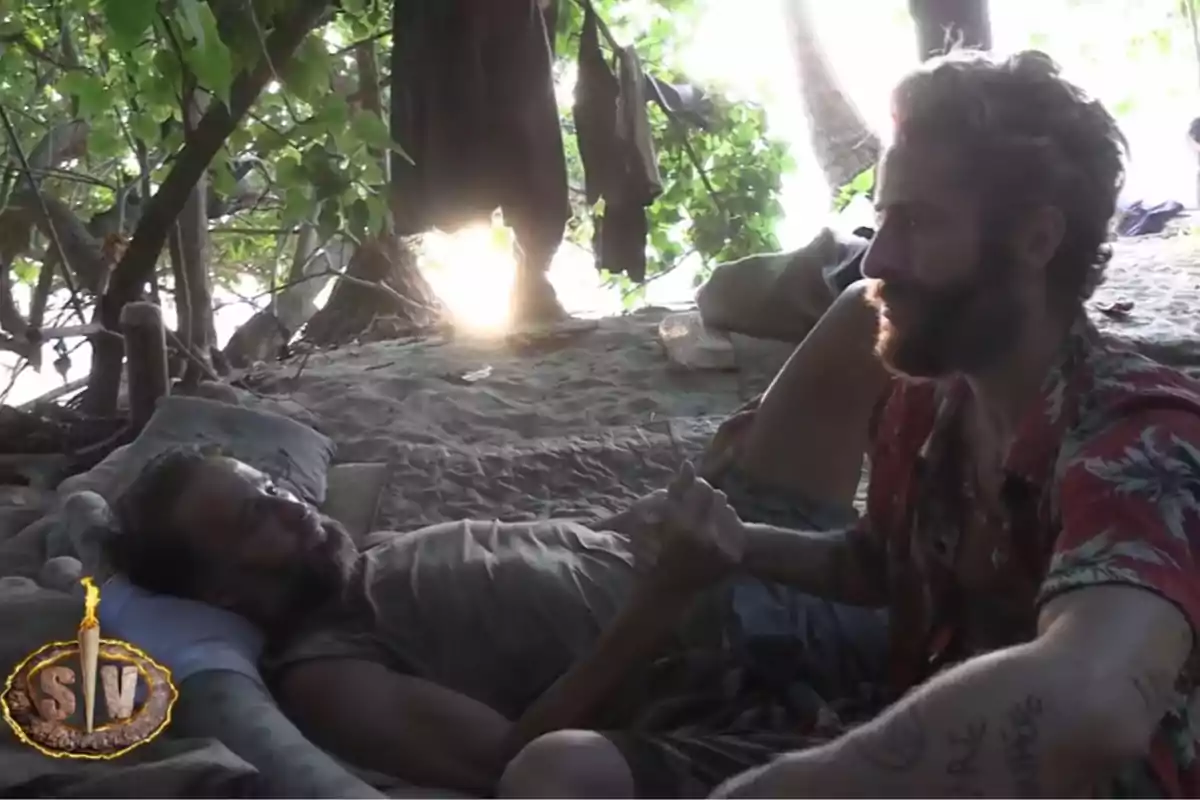 Álvaro Muñoz Escassi y Pelayo Díaz se dan la mano en una playa bajo la sombra de los árboles, uno está acostado y el otro sentado, con luz de sol filtrándose entre las hojas y ropa colgada al fondo. Captura de 'Supervivientes' Álvaro Muñoz Escassi y Pelayo Díaz se dan la mano en una playa bajo la sombra de los árboles, uno está acostado y el otro sentado, con luz de sol filtrándose entre las hojas y ropa colgada al fondo. Captura de 'Supervivientes'