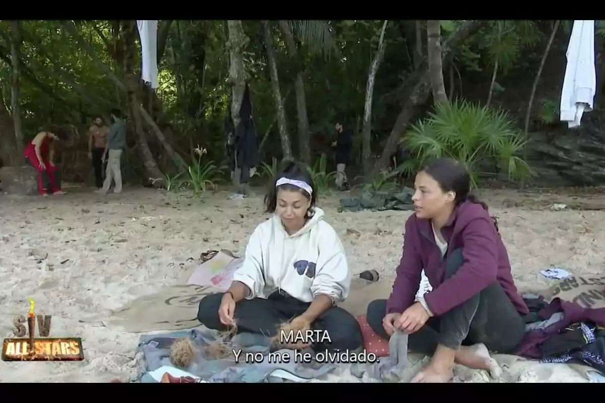 Lola Mencía y Marta Peñate sentadas en la arena de una playa, rodeadas de vegetación, con subtítulos y el logotipo de 'Supervivientes'. Captura del 18 de julio de 2024