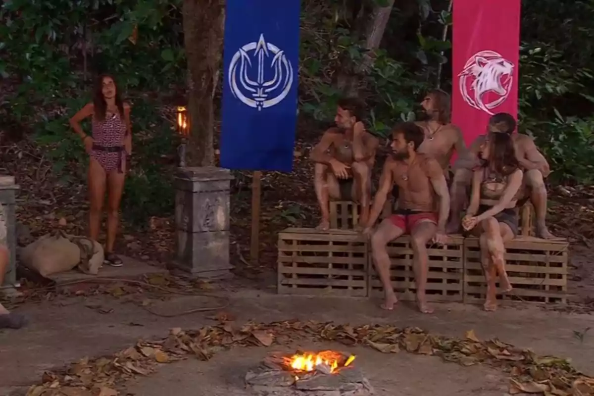 Un grupo de personas sentadas en un entorno selvático alrededor de una fogata, con banderas de colores azul y rojo en el fondo. Captura de 'Supervivientes' Un grupo de personas sentadas en un entorno selvático alrededor de una fogata, con banderas de colores azul y rojo en el fondo. Captura de 'Supervivientes'