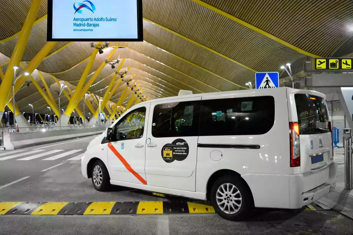 A white taxi van is parked outside Adolfo Suárez Madrid-Barajas Airport, with a modern architectural design and yellow columns visible in the background.