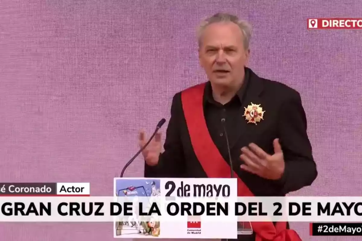 José Coronado con una banda roja y una medalla en el pecho habla en un podio durante un evento del 2 de mayo. José Coronado con una banda roja y una medalla en el pecho habla en un podio durante un evento del 2 de mayo.