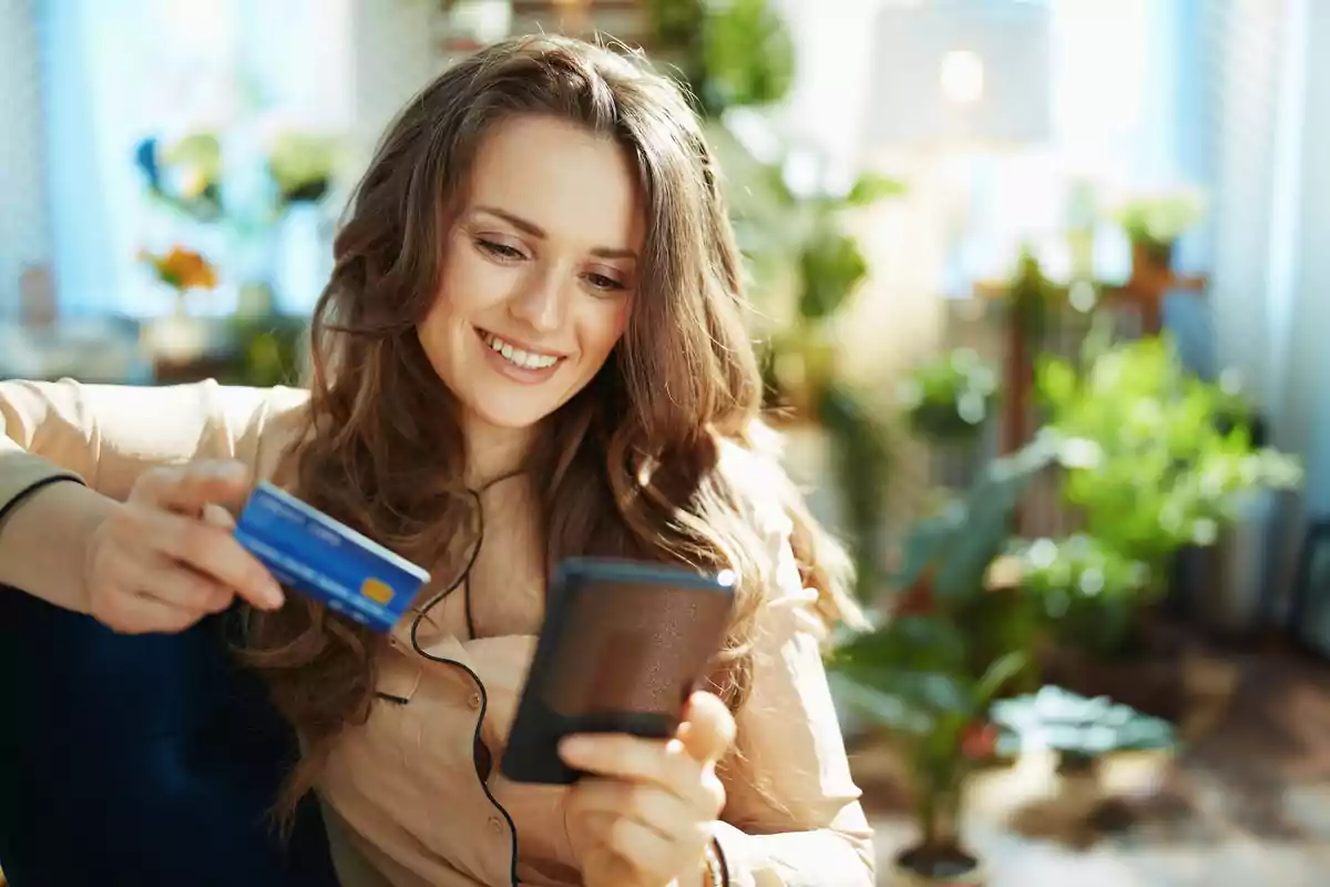 Mujer sonriendo mientras sostiene una tarjeta de crédito y un teléfono móvil en un entorno hogareño con plantas.