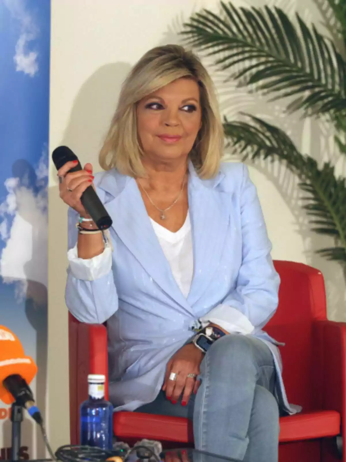 Terelu Campos sentada en una silla roja sosteniendo un micrófono, vestida con chaqueta azul claro y pantalón de mezclilla, con fondo de planta y micrófonos sobre la mesa. Terelu Campos sentada en una silla roja sosteniendo un micrófono, vestida con chaqueta azul claro y pantalón de mezclilla, con fondo de planta y micrófonos sobre la mesa.
