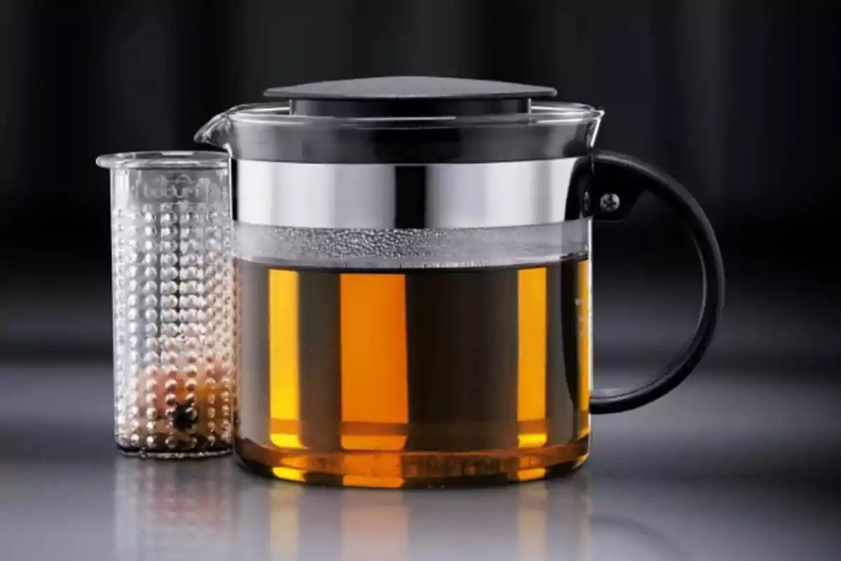 Tetera de vidrio con infusor de metal y asa negra sobre una superficie gris Tetera de vidrio con infusor de metal y asa negra sobre una superficie gris