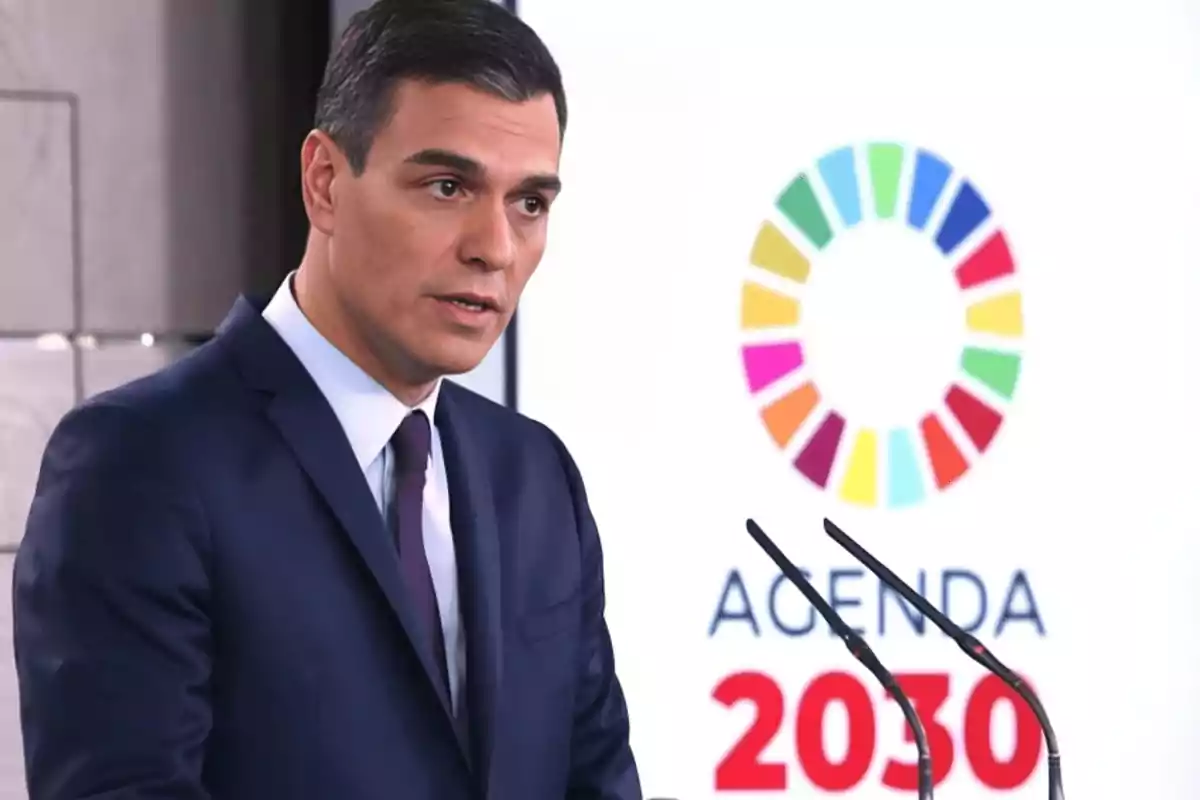 Un hombre en traje habla frente a un micrófono con un cartel de la Agenda 2030 de fondo.