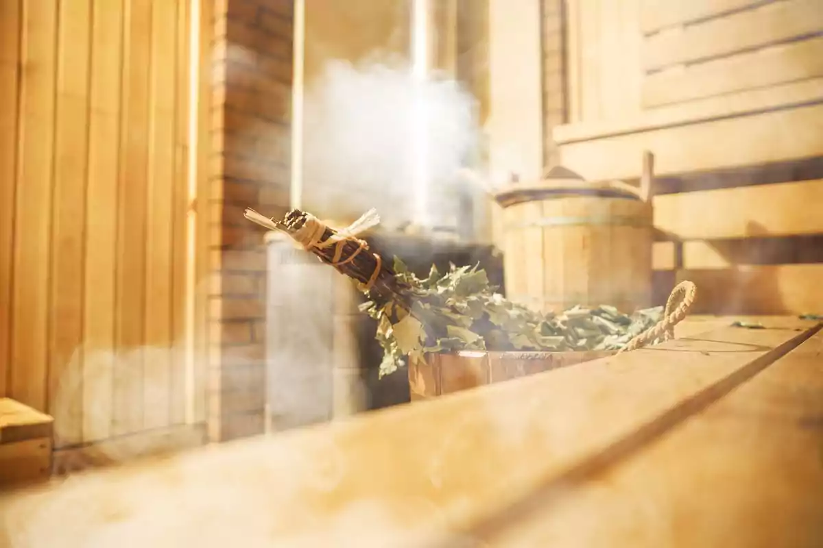 Una sauna de madera con un balde y un ramo de hojas sobre un banco, rodeada de vapor.