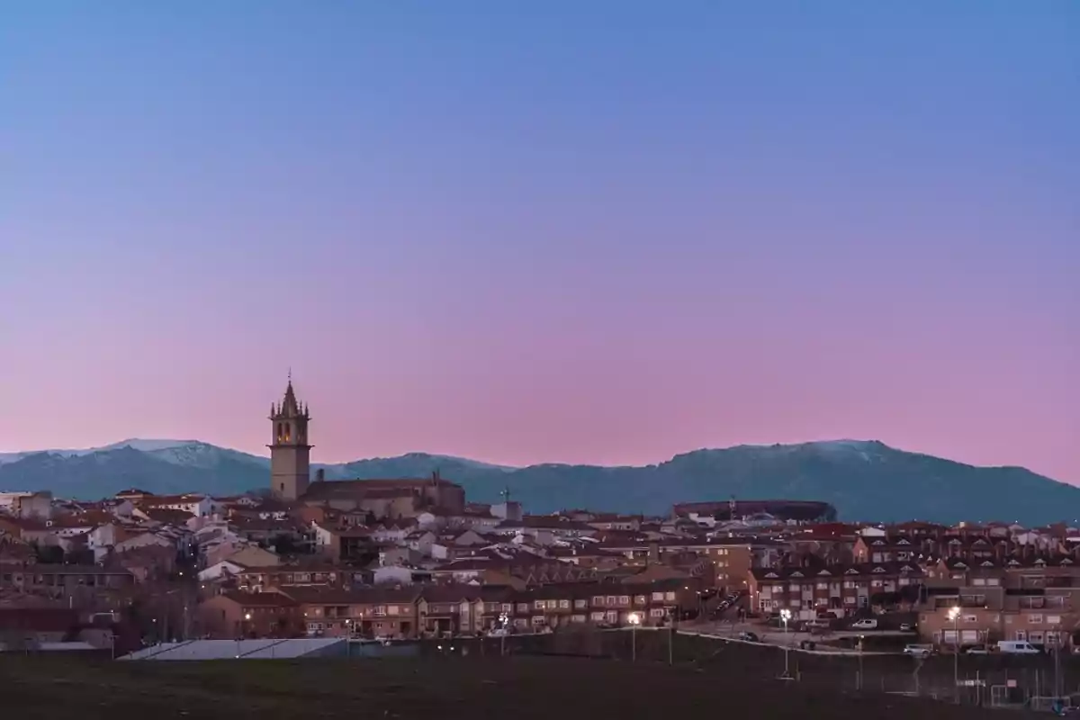 Vista de un pueblo con una iglesia destacada al atardecer y montañas al fondo.