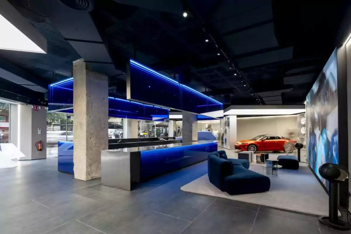 Una sala de exhibición moderna con un diseño futurista, que incluye un coche deportivo rojo, muebles elegantes y una iluminación azul vibrante.