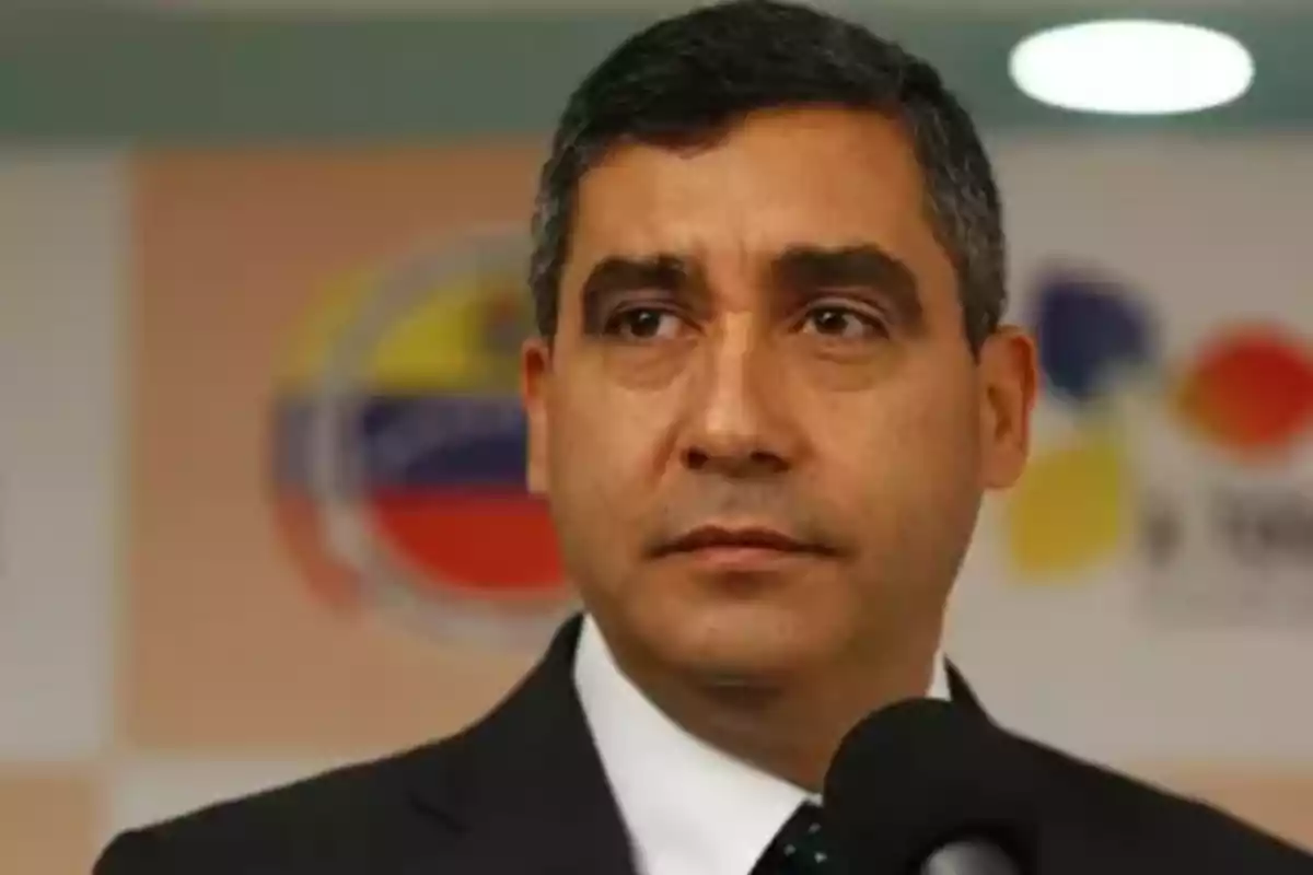Un hombre de traje oscuro y corbata habla frente a un micrófono con un fondo borroso que incluye un logotipo colorido. Un hombre de traje oscuro y corbata habla frente a un micrófono con un fondo borroso que incluye un logotipo colorido.