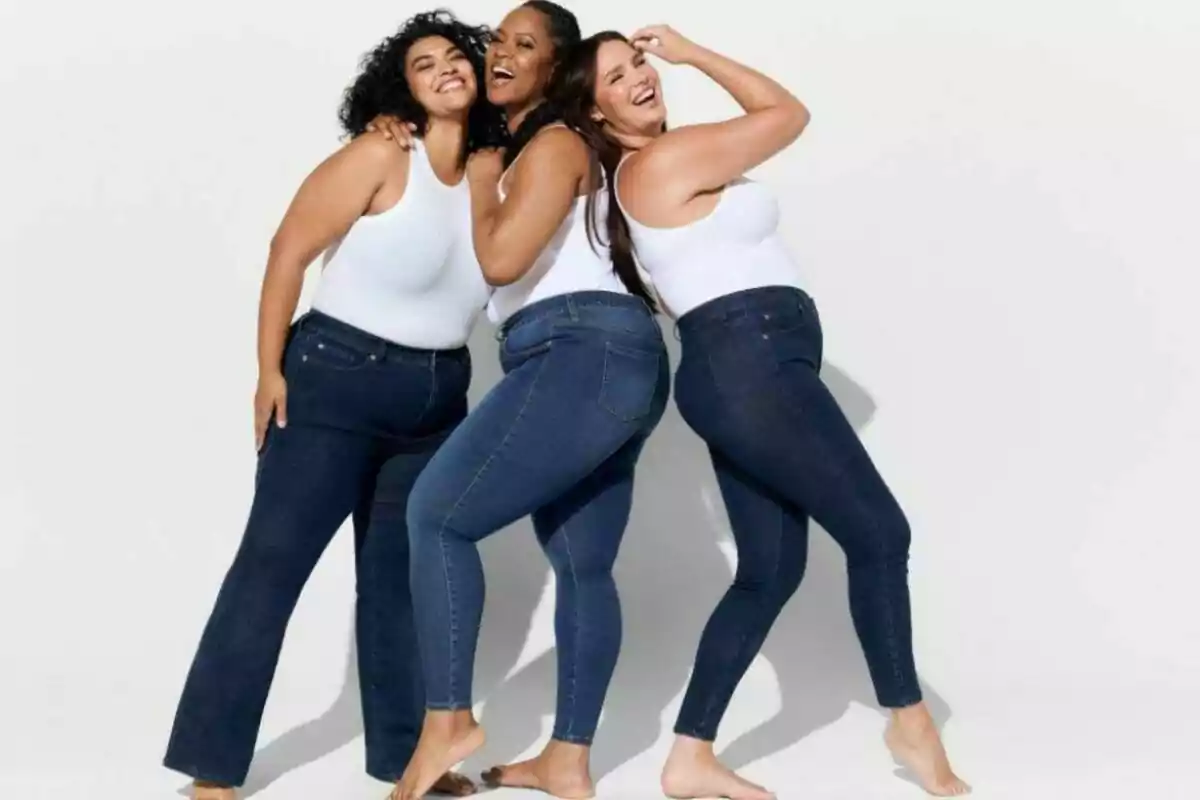 Three smiling women pose together wearing white sleeveless jerseys and fitted denim jeans, barefoot on a light background. Three smiling women pose together wearing white sleeveless jerseys and fitted denim jeans, barefoot on a light background.