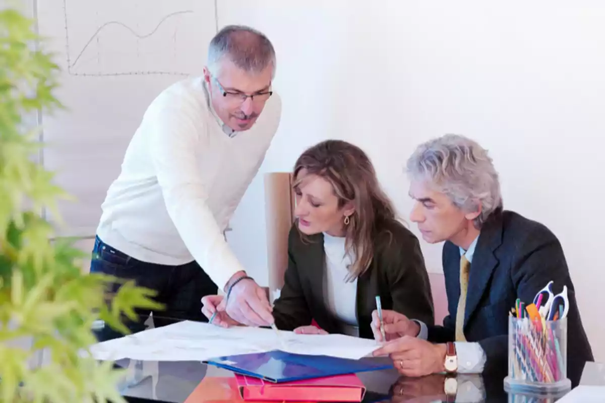 Tres personas en una reunión de trabajo revisando documentos en una mesa con gráficos en el fondo. Tres personas en una reunión de trabajo revisando documentos en una mesa con gráficos en el fondo.