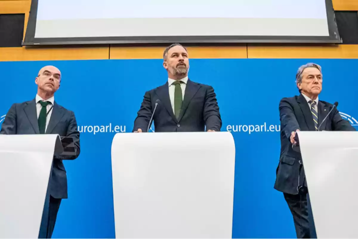 Tres hombres de pie detrás de podios blancos en una conferencia de prensa con un fondo azul que tiene el texto "europarl.eu".