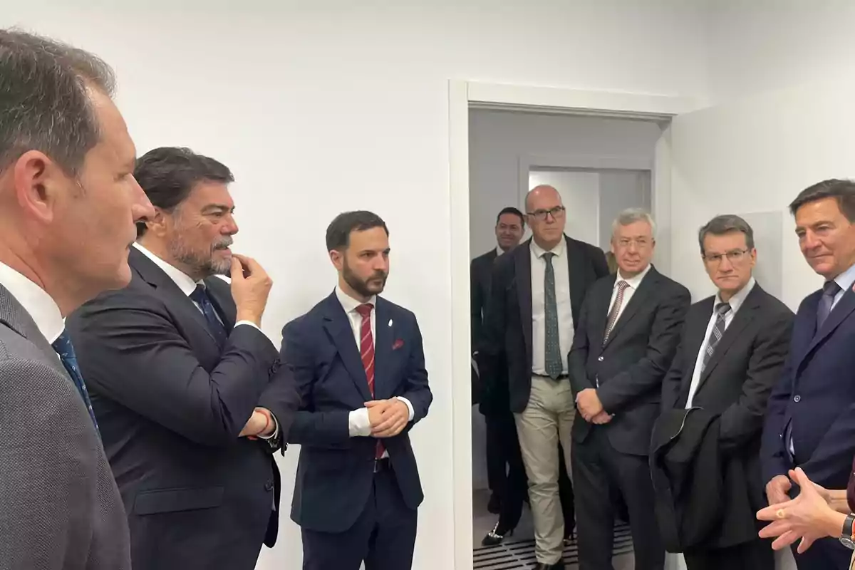 A group of men in formal suits is gathered in a white room, some of them seem to be conversing while others are observing.