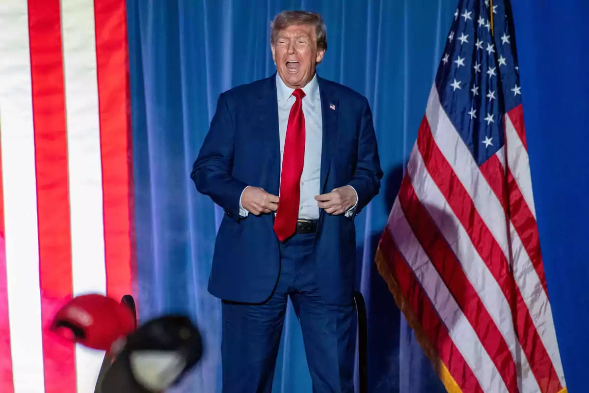A man in a blue suit and red tie stands in front of an American flag and a blue background. A man in a blue suit and red tie stands in front of an American flag and a blue background.