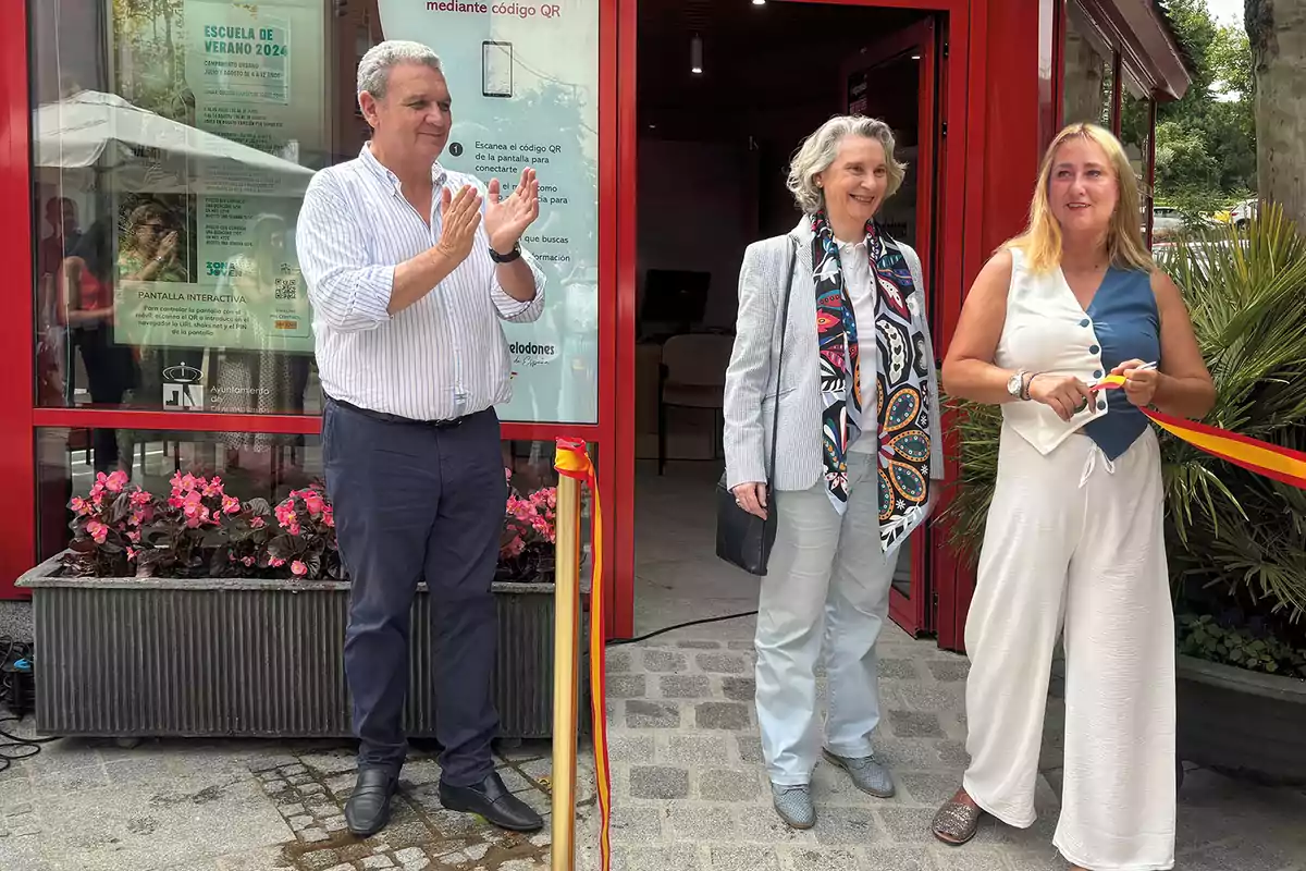 Tres personas están en una ceremonia de inauguración, una de ellas está cortando una cinta mientras otra aplaude y la tercera sonríe, con un fondo de flores y un cartel que anuncia una escuela de verano.