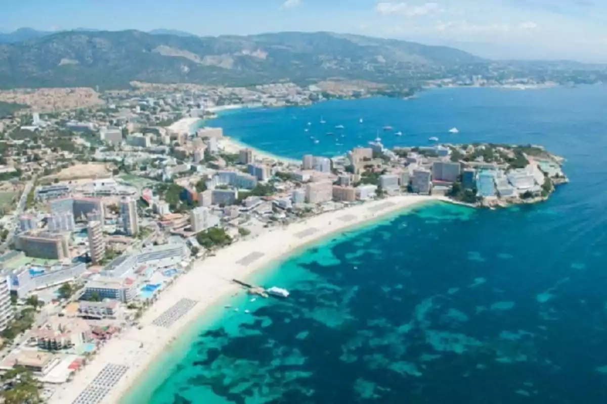 Vista aérea de una playa con edificios y montañas al fondo en un día soleado.