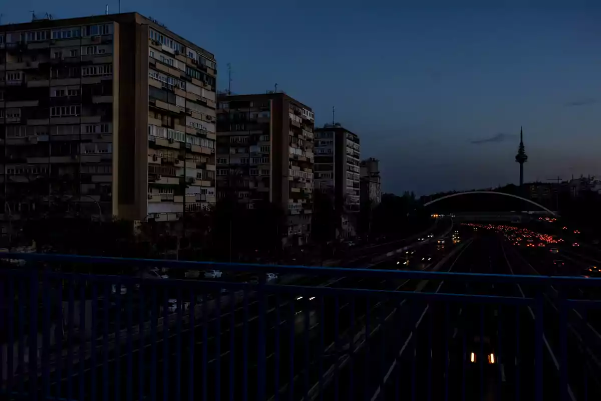 Una vista nocturna de una ciudad con edificios altos a la izquierda y una carretera iluminada por las luces de los coches a la derecha, con una torre visible en el horizonte. Una vista nocturna de una ciudad con edificios altos a la izquierda y una carretera iluminada por las luces de los coches a la derecha, con una torre visible en el horizonte.