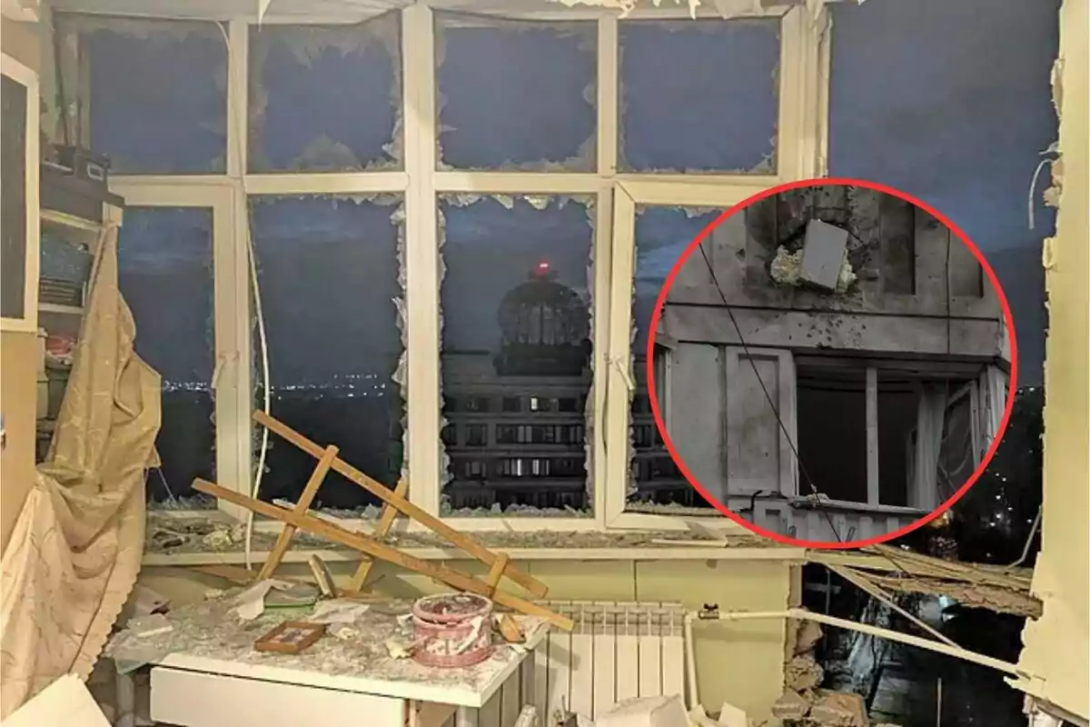 Una habitación con una ventana rota y escombros en el interior, con un edificio visible a través de la ventana y un detalle circular que muestra un daño en la fachada exterior.