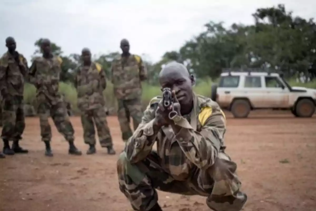 Un grupo de soldados en uniforme militar camuflado, uno de ellos en primer plano apuntando con un arma, con un vehículo todoterreno al fondo.