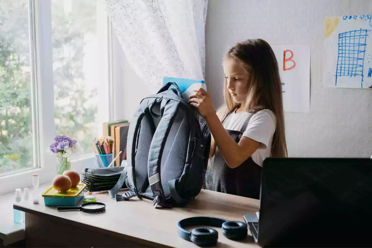 Una niña empacando su mochila en un escritorio con útiles escolares, una computadora portátil y una ventana al fondo.