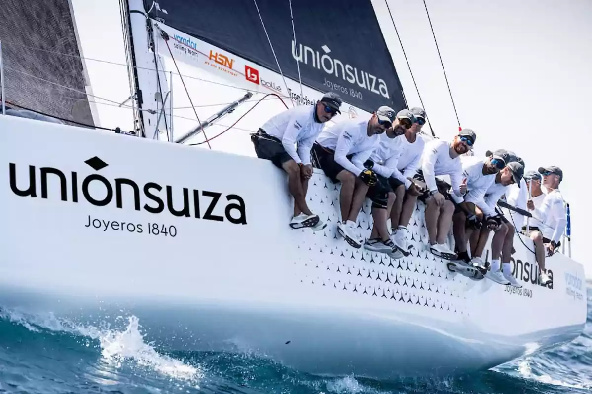 Un grupo de personas con ropa deportiva blanca y gorras está sentado en el borde de un velero blanco con la inscripción "unión suiza Joyeros 1840" mientras navegan en el mar.