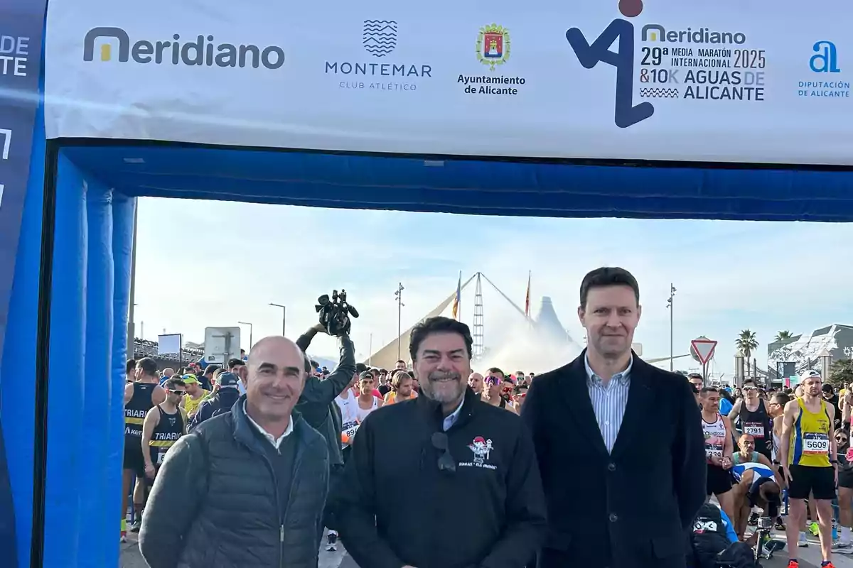 Three men pose in front of an inflatable arch at a marathon event in Alicante with runners and sponsor logos visible.