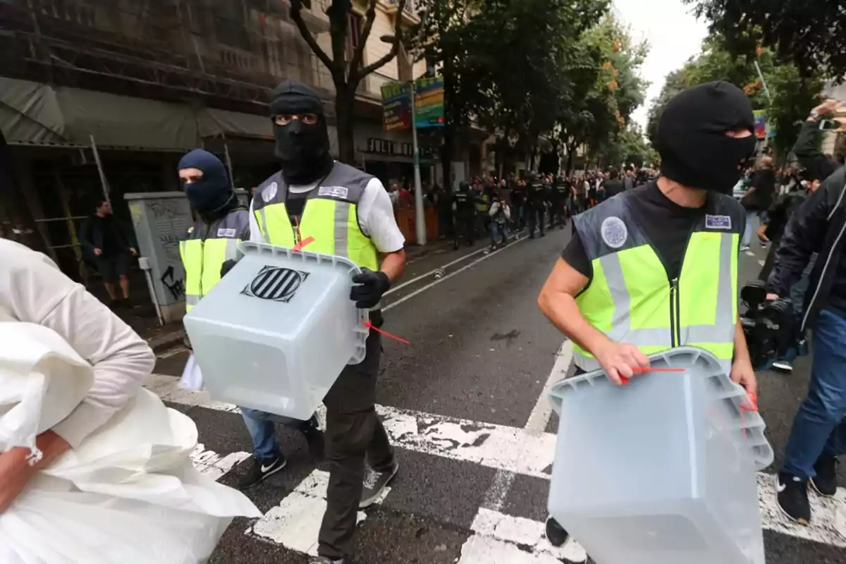 Agentes con chalecos reflectantes y pasamontañas transportan urnas de plástico transparentes en una calle rodeada de personas y árboles Agentes con chalecos reflectantes y pasamontañas transportan urnas de plástico transparentes en una calle rodeada de personas y árboles