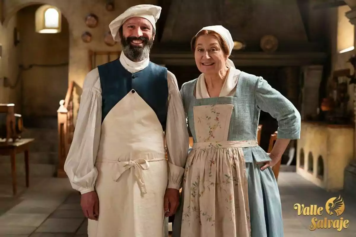 Amadeo y Eva vestida con ropa de época, ambos sonrientes, en el interior de una cocina antigua con utensilios de cocina en el fondo. Amadeo y Eva vestida con ropa de época, ambos sonrientes, en el interior de una cocina antigua con utensilios de cocina en el fondo.