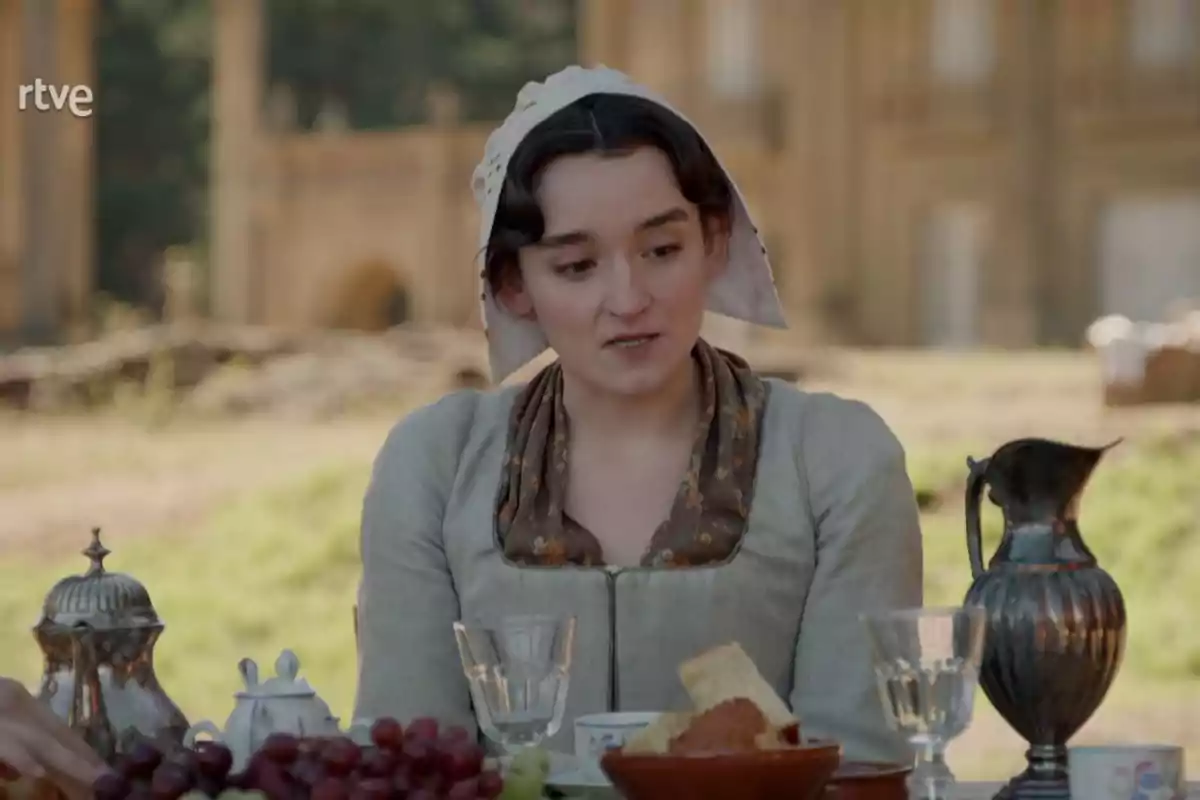 Matilde dressed in period clothing and a headscarf is sitting at an outdoor table with food and drinks, in a rural setting with an old building in the background. Still from 'Valle Salvaje'