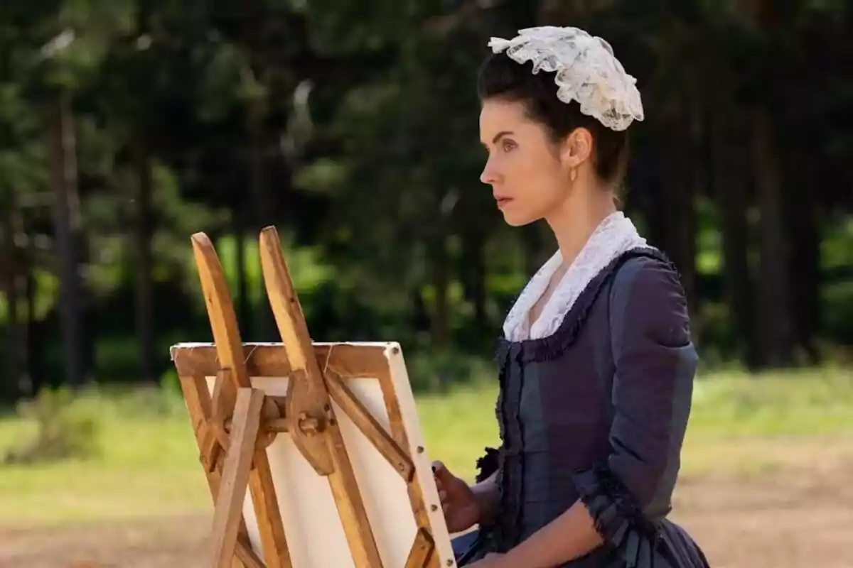 Mercedes vestida con ropa de época y un tocado blanco está sentada al aire libre frente a un caballete de pintura en un entorno natural. Captura de 'Valle Salvaje'