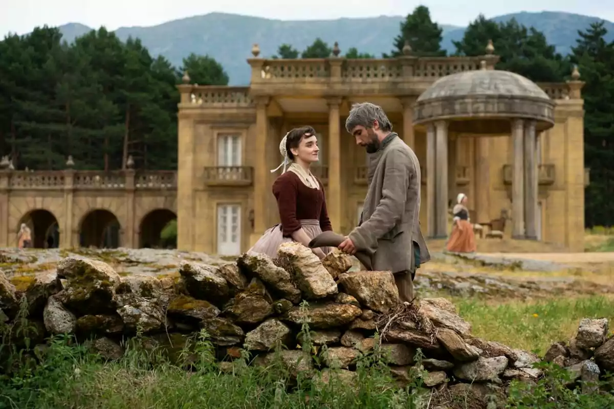 Dos personas vestidas con ropa de época conversan junto a un muro de piedra, con un edificio antiguo y árboles en el fondo en la serie Valle Salvaje.