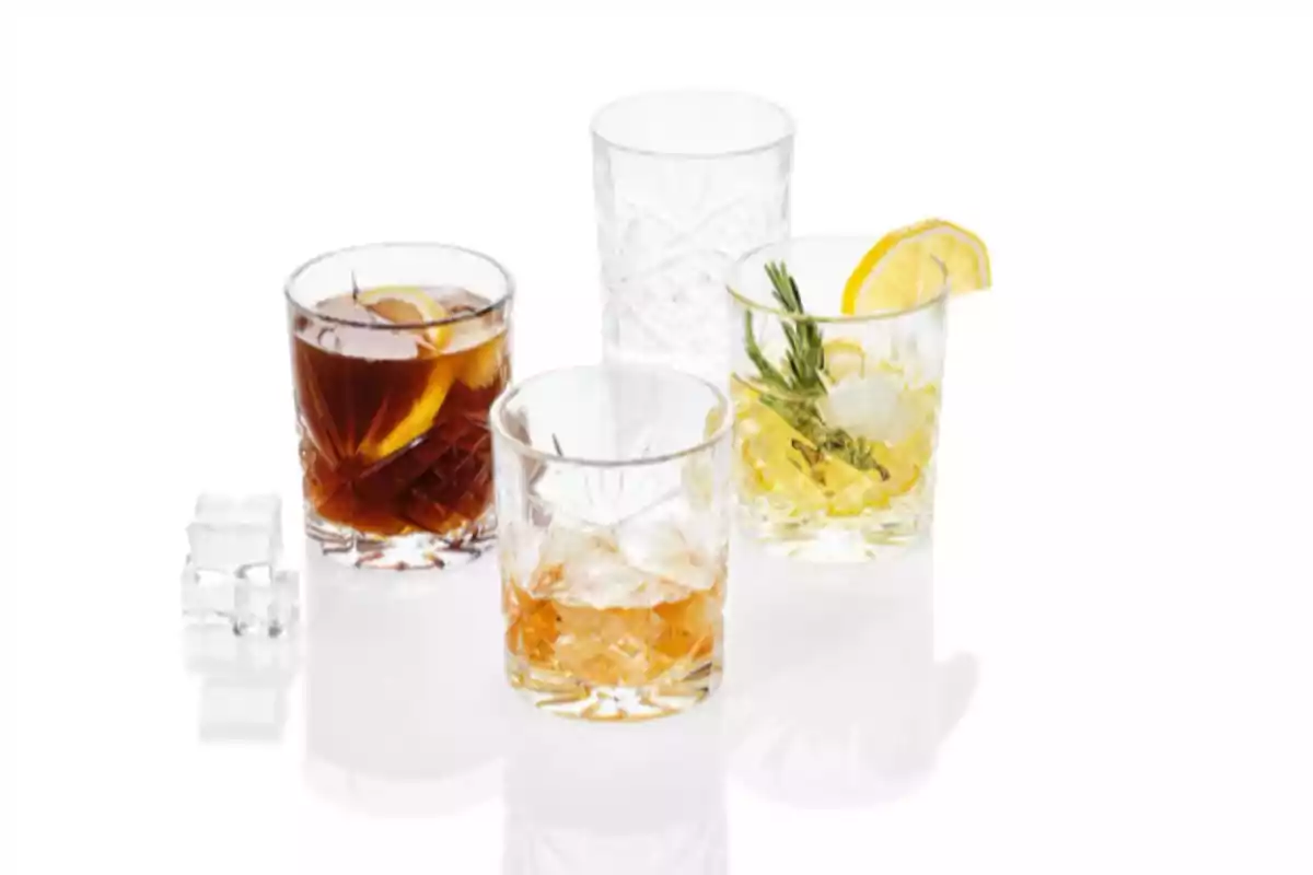 Cuatro vasos de cristal con diferentes bebidas y hielo, uno decorado con rodaja de limón y ramita de romero, sobre fondo blanco Cuatro vasos de cristal con diferentes bebidas y hielo, uno decorado con rodaja de limón y ramita de romero, sobre fondo blanco
