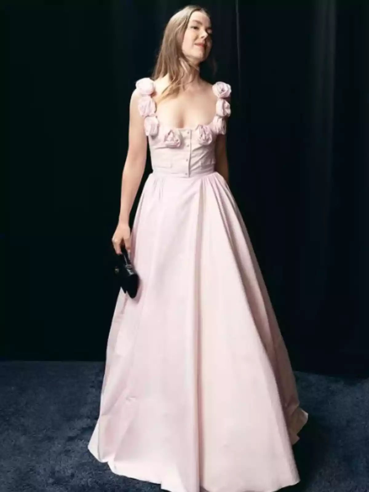 Mujer con vestido largo rosa claro decorado con flores en los tirantes y sosteniendo un bolso negro sobre fondo oscuro Mujer con vestido largo rosa claro decorado con flores en los tirantes y sosteniendo un bolso negro sobre fondo oscuro