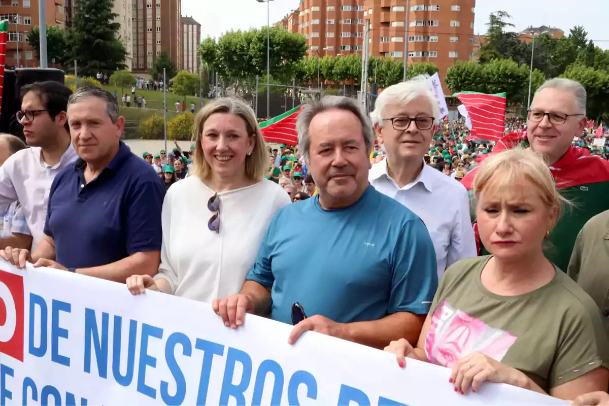 Un grupo de personas sostiene una pancarta durante una manifestación al aire libre con edificios y árboles al fondo y una multitud participando en la protesta