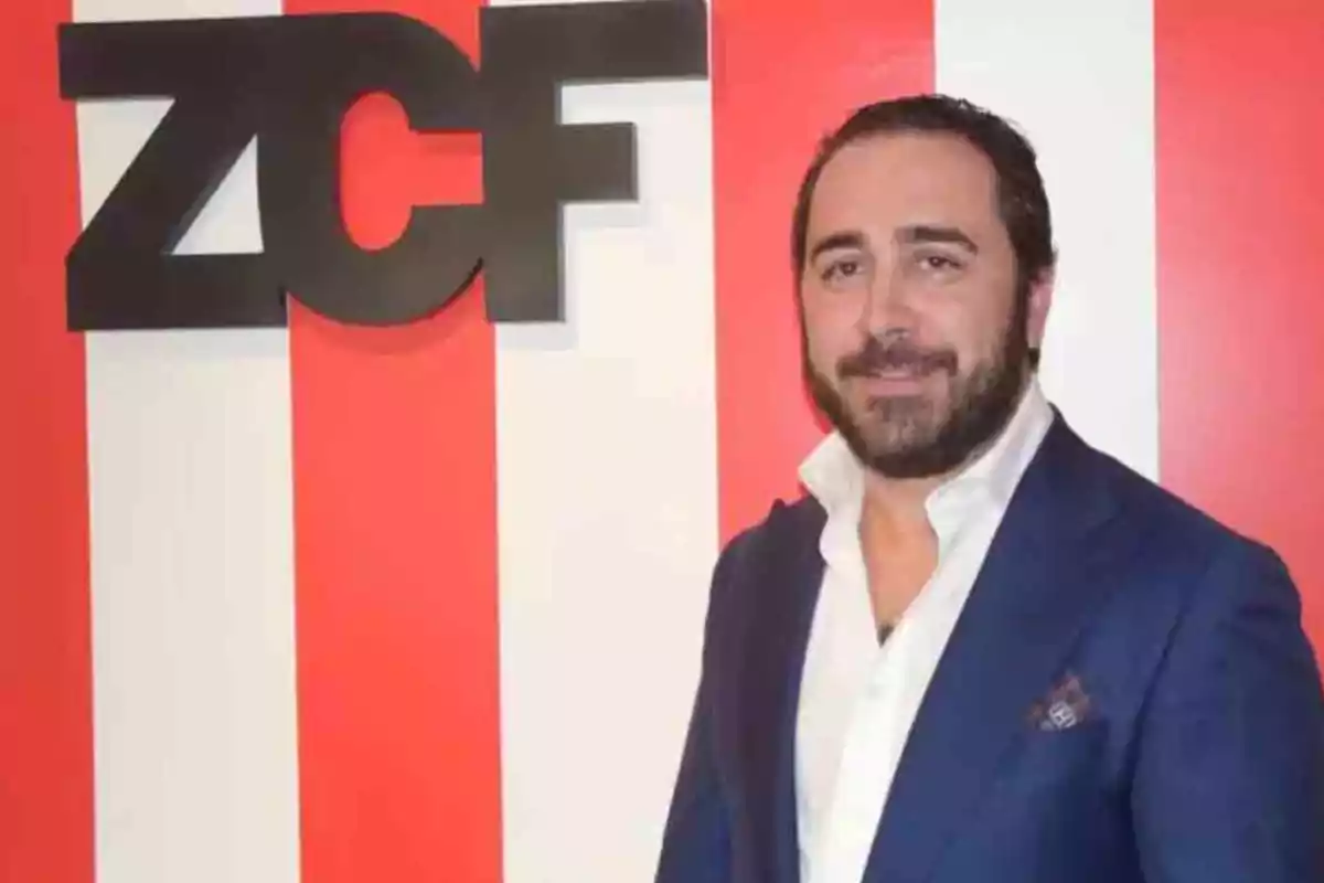 Hombre con barba y saco azul posando frente a una pared con franjas rojas y blancas y un letrero que dice ZCF Hombre con barba y saco azul posando frente a una pared con franjas rojas y blancas y un letrero que dice ZCF
