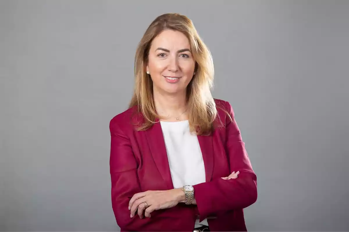 Mujer con chaqueta roja y blusa blanca sonriendo con los brazos cruzados frente a un fondo gris.