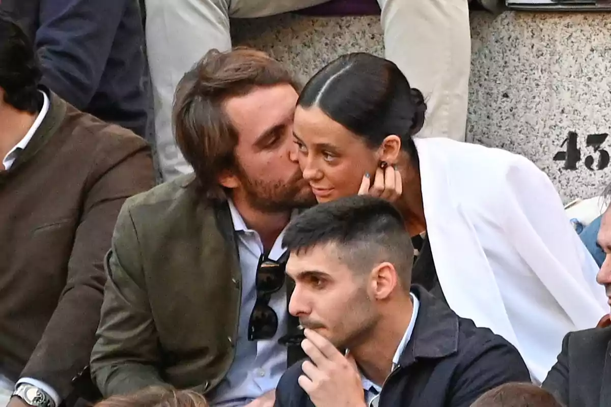 A couple sitting at an event, where a man kisses a woman on the cheek who smiles, while other people are around. A couple sitting at an event, where a man kisses a woman on the cheek who smiles, while other people are around.