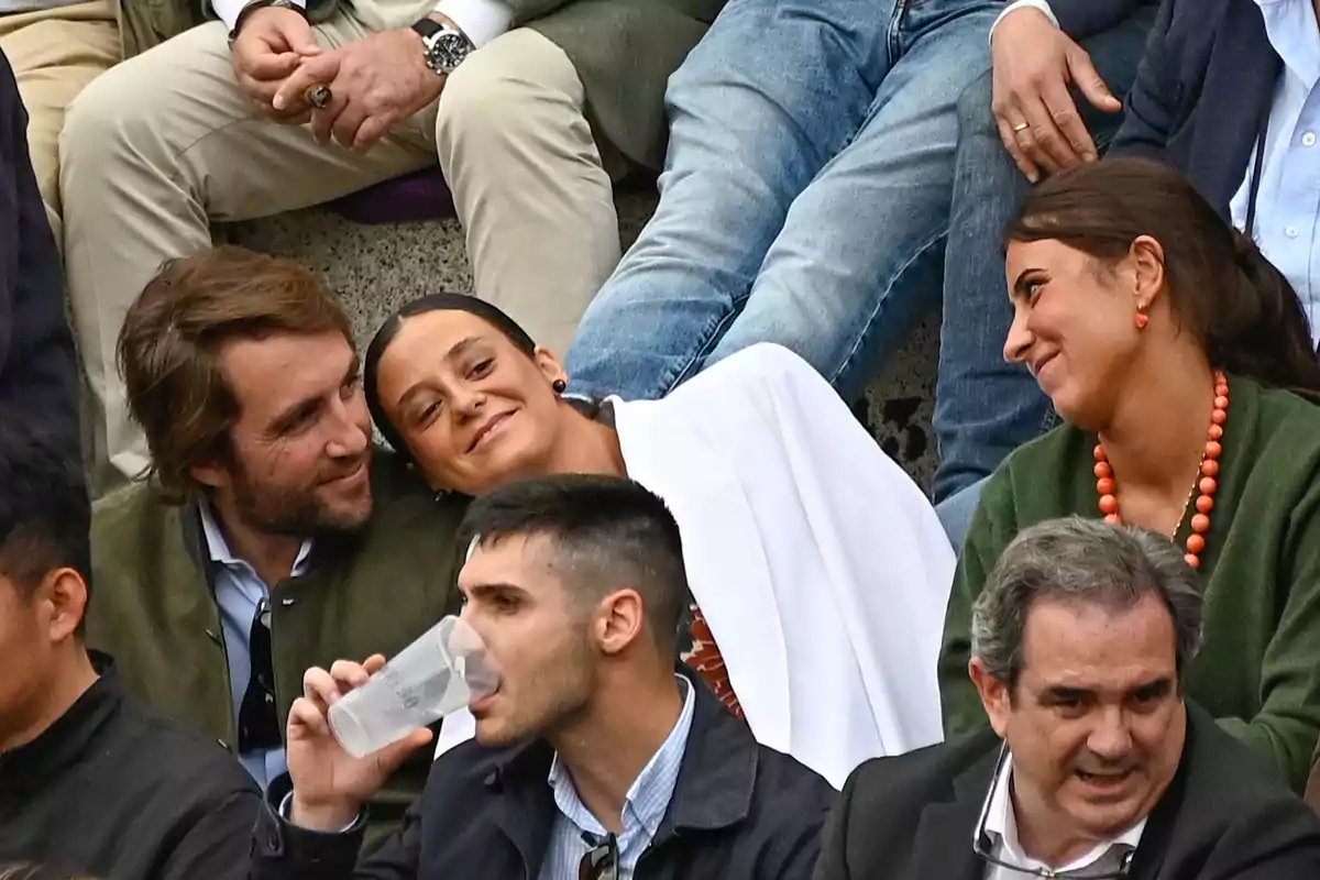A group of people sitting in bleachers, some talking and smiling, while one person drinks from a cup. A group of people sitting in bleachers, some talking and smiling, while one person drinks from a cup.