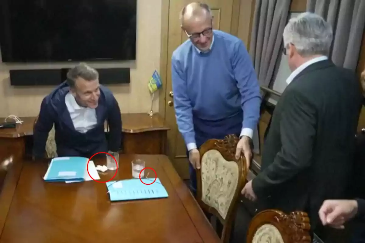 Tres personas en una sala de reuniones, dos de ellas de pie y una inclinándose hacia adelante, con documentos y vasos sobre la mesa. Tres personas en una sala de reuniones, dos de ellas de pie y una inclinándose hacia adelante, con documentos y vasos sobre la mesa.