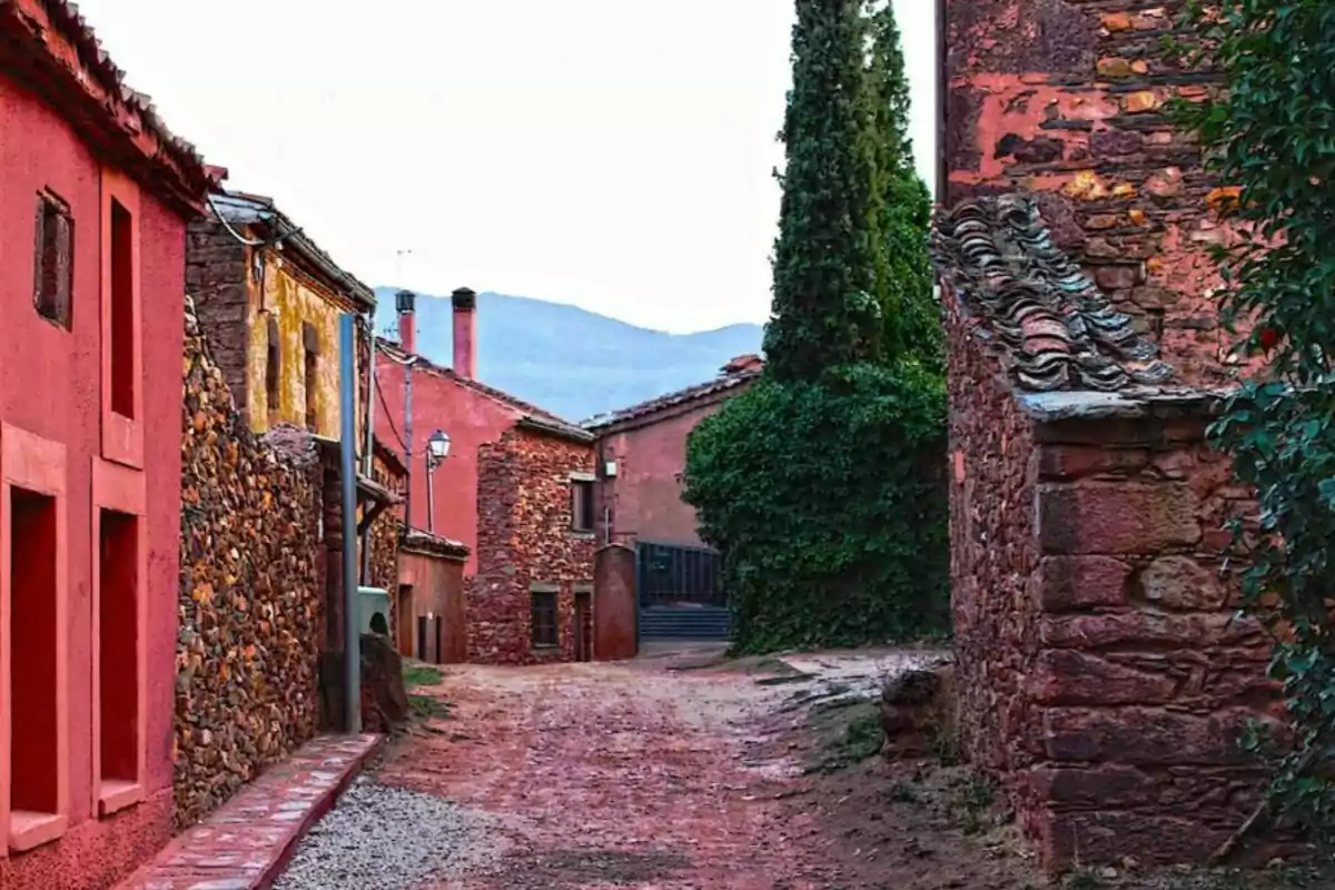 Callejón de un pueblo con casas de piedra y fachadas de color rojo, rodeado de vegetación y montañas al fondo.