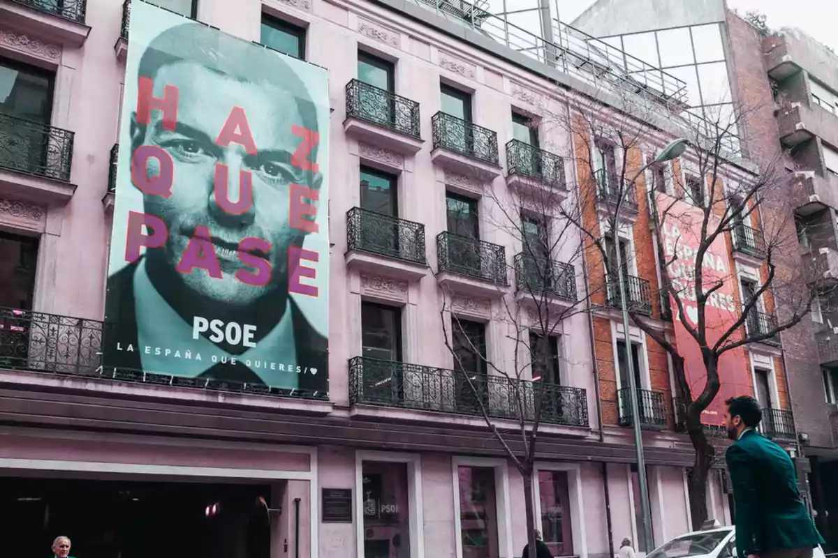 Fachada de edificio con carteles de campaña política del PSOE y personas caminando por la acera