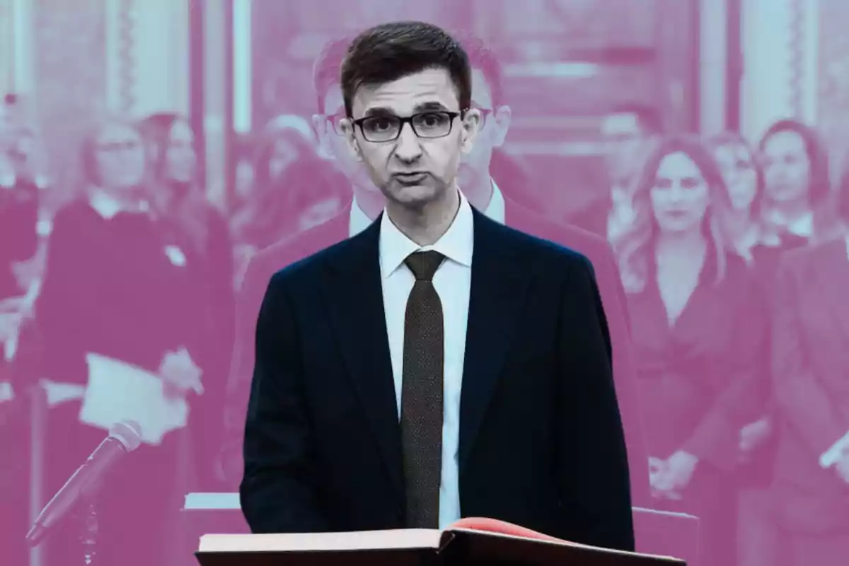 Un hombre con gafas y traje oscuro está de pie frente a un micrófono y un libro abierto, con un fondo de personas desenfocadas en tonos rosados.