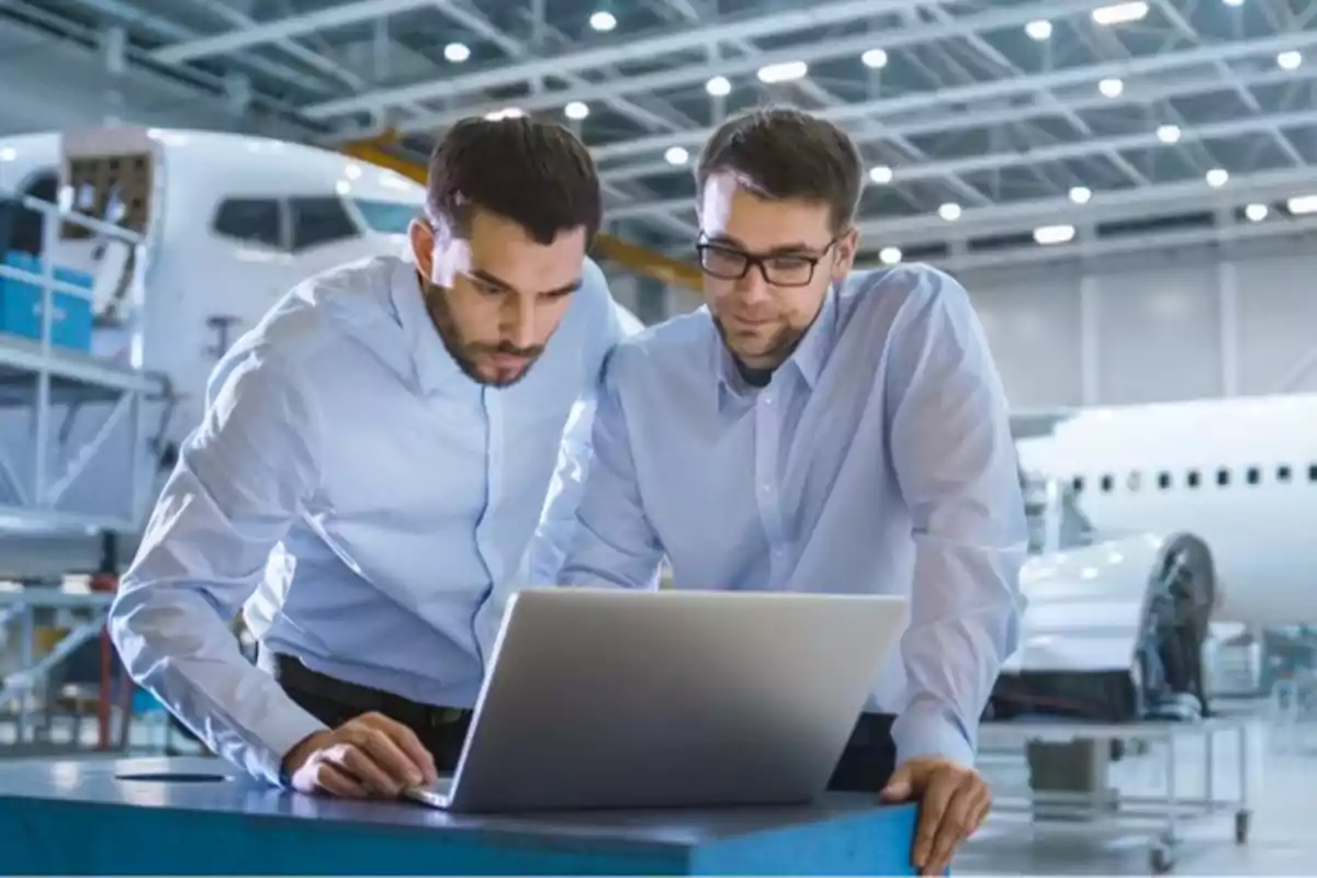 Dos ingenieros revisando información en una computadora portátil dentro de un hangar de aviones.