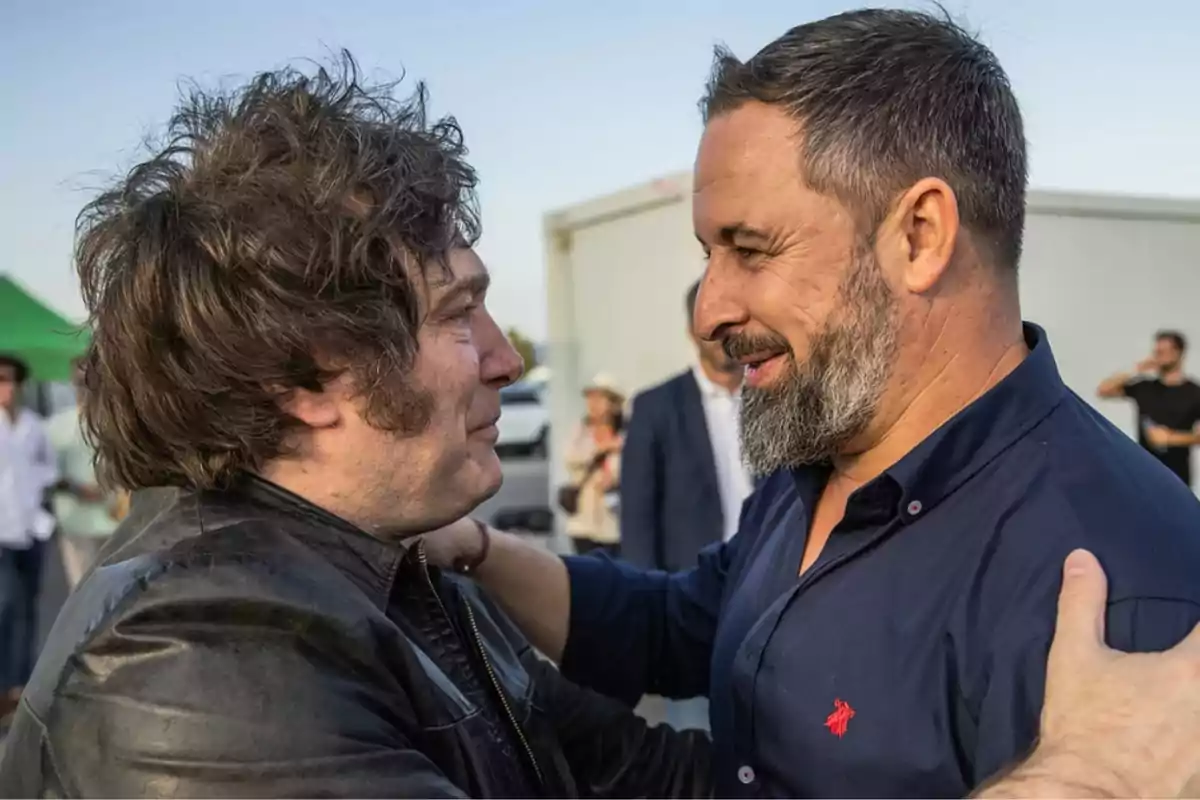 Two men greet each other and smile as they hug, with people and structures blurred in the background.