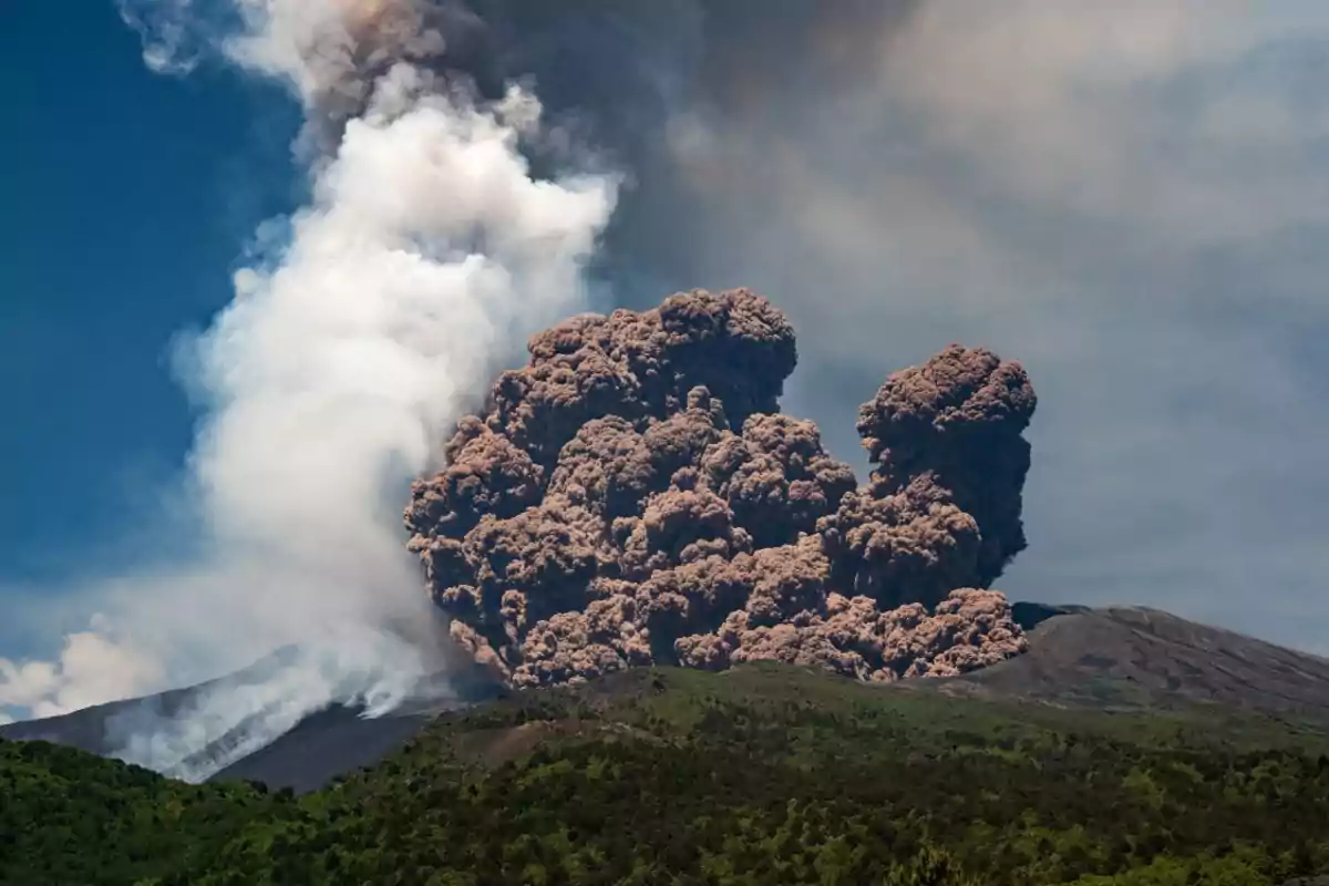 Erupción volcánica con una gran nube de ceniza y humo elevándose sobre un paisaje verde bajo un cielo azul Erupción volcánica con una gran nube de ceniza y humo elevándose sobre un paisaje verde bajo un cielo azul